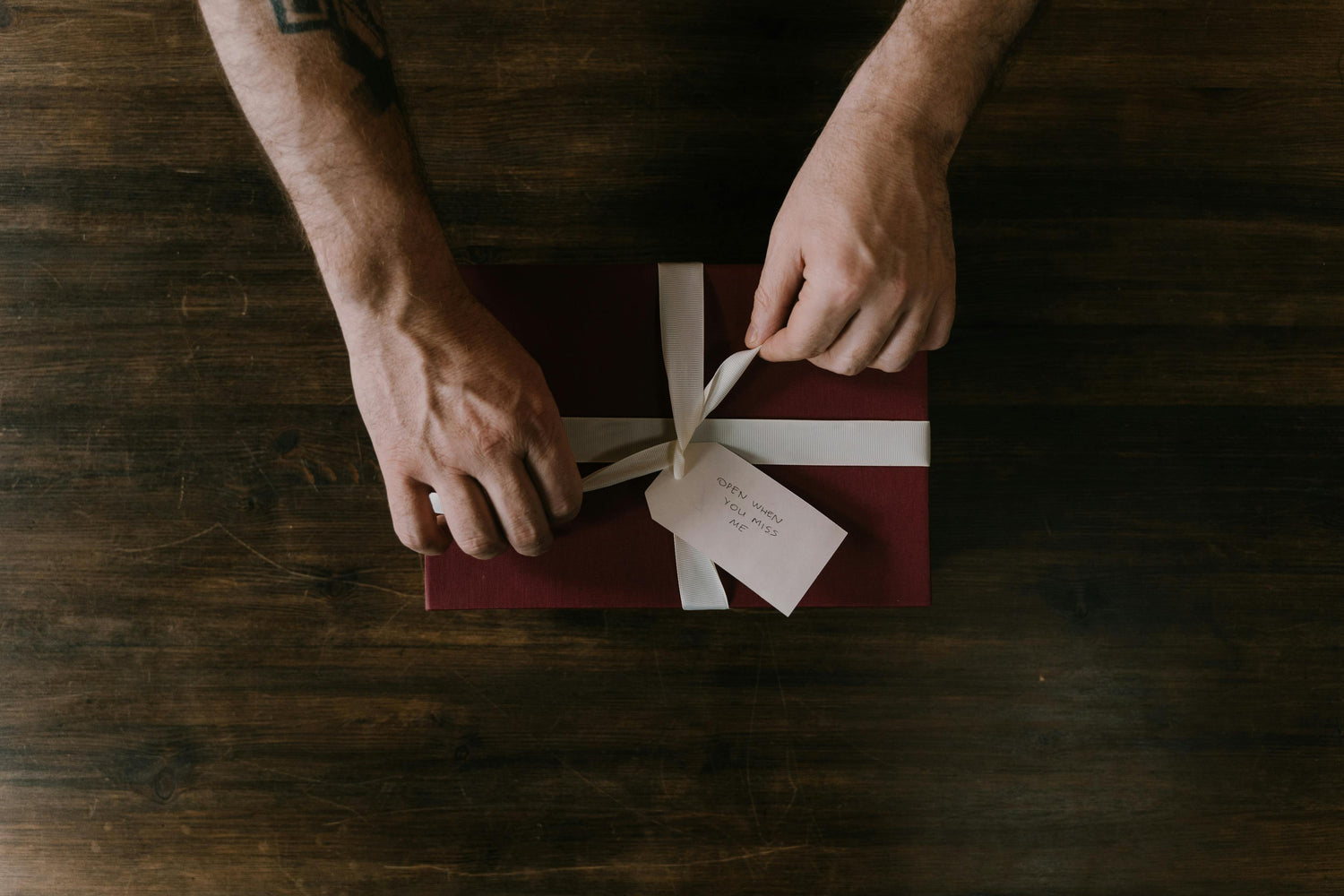 A pair of hands tying a bow on a red package with a tag that reads "open when you miss me" on the package