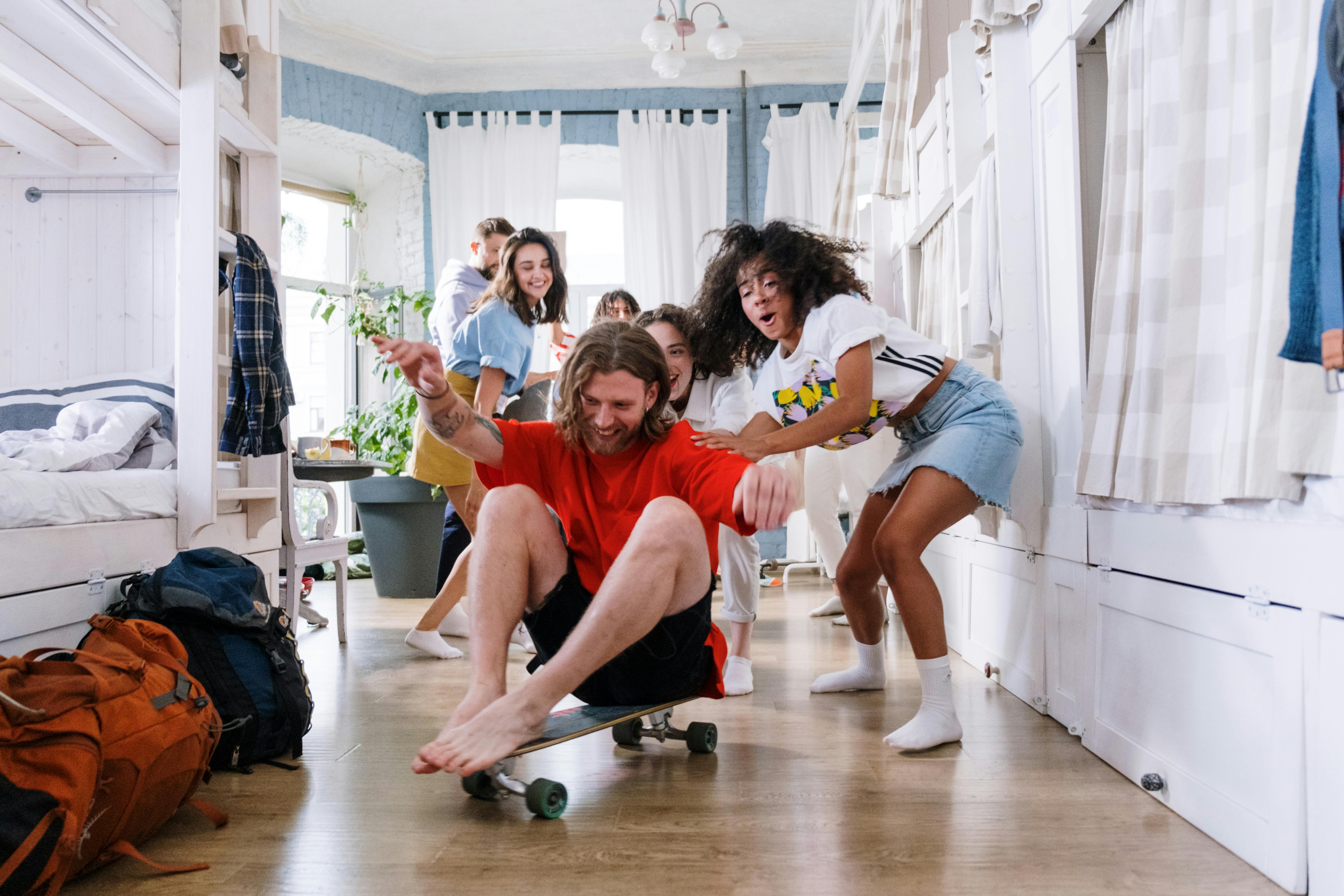 A bunch of young adult friends playing around in a blue-painted room with large windows behind them. A young man is being pushed around on the floor, laughing, on a skateboard.